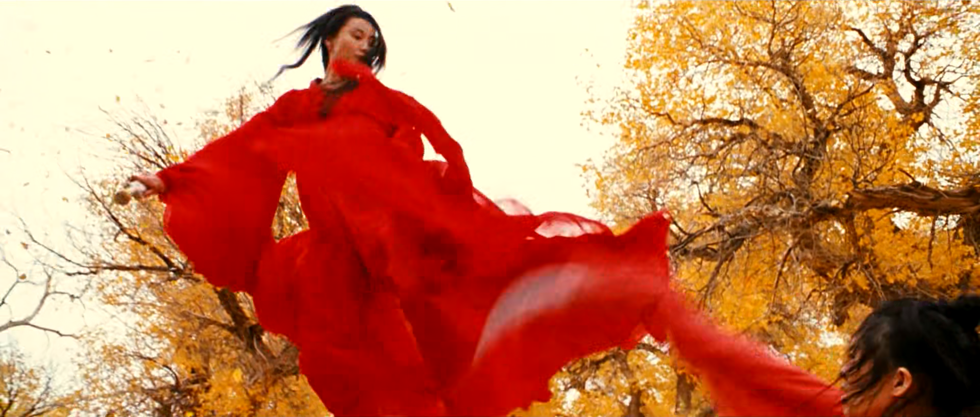 A red-robed figure whirls a broad sheet of red fabric beneath a yellow-leaved tree and pale sky.