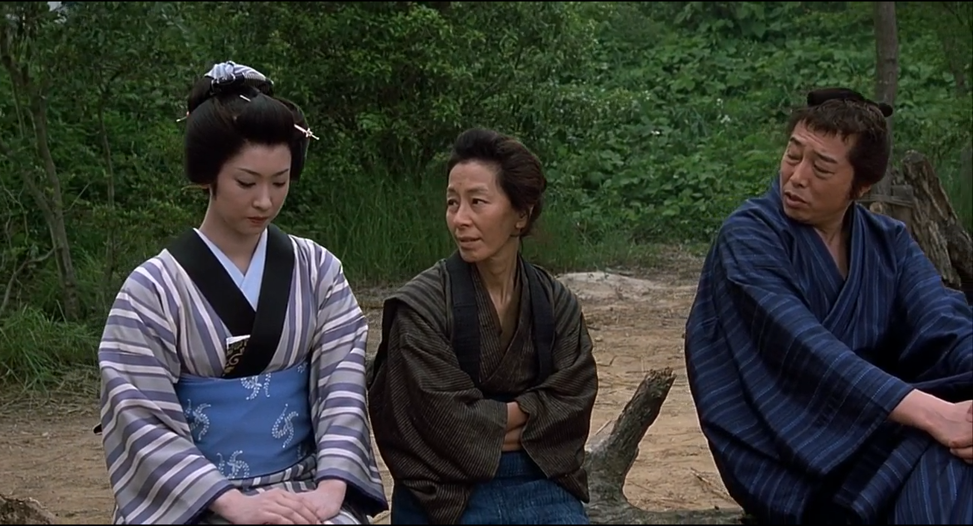 Three people in muted blue, black and striped robes sit outdoors against dense green foliage.