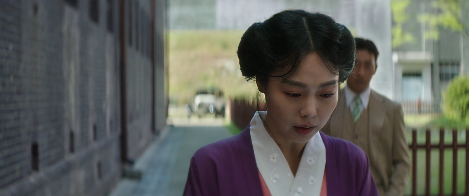 A woman in a purple robe walks with her gaze lowered as suited figures follow through a grey courtyard.