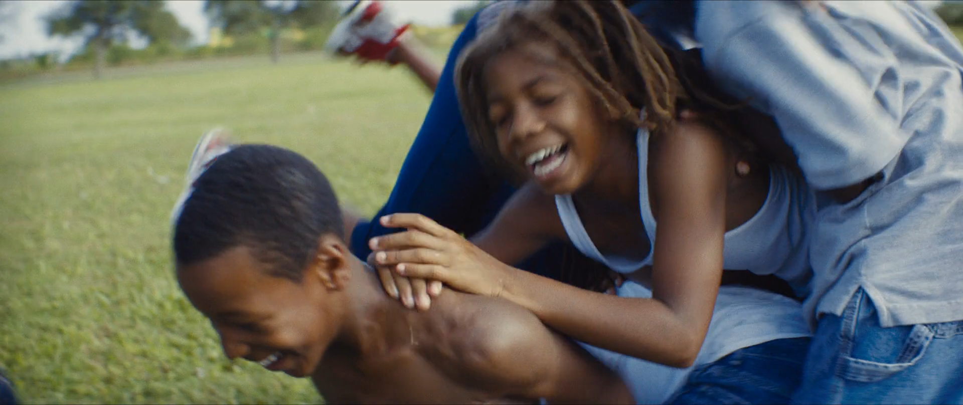 Children in blue and white play-wrestle on sunlit green grass, warm skin against the blue of their clothes.