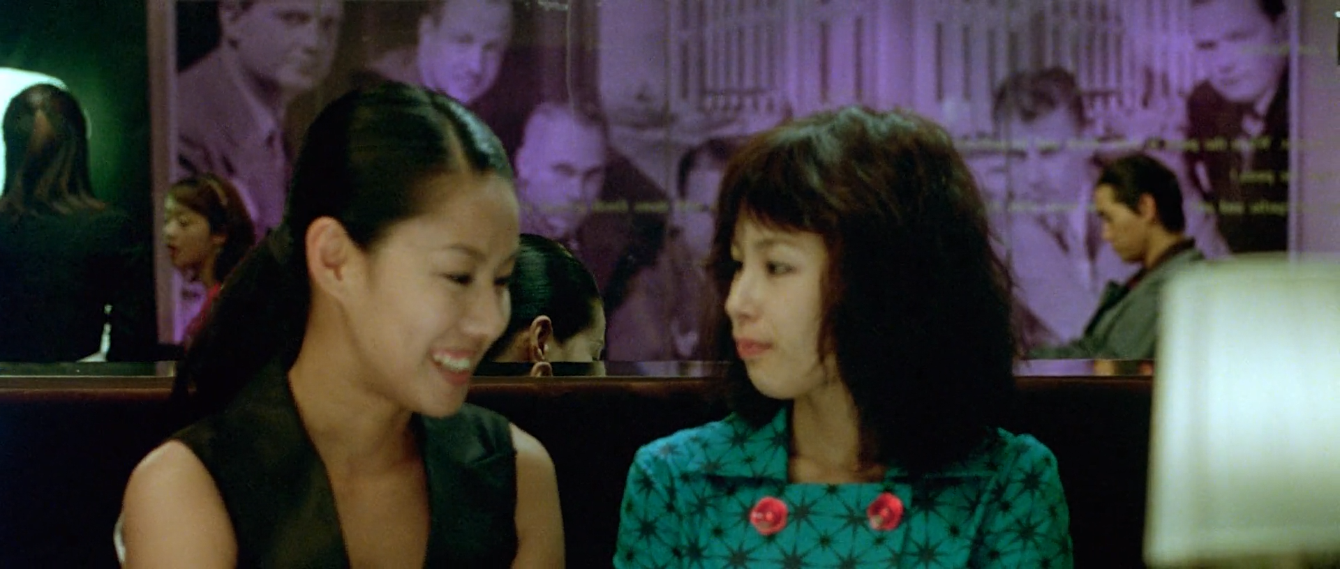 Two women sit talking in a dim club, one in black and one in a teal patterned dress with red buttons, against a violet photographic mural.