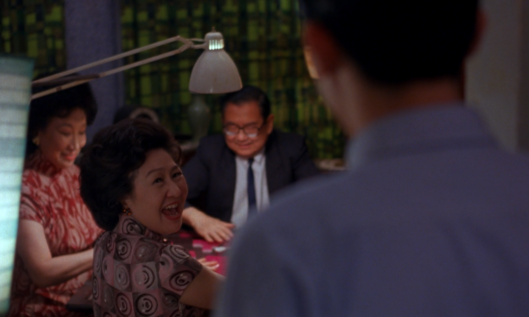 A woman in a patterned dress laughs at a mahjong table under a domed lamp, with a blue-grey shoulder in the foreground and a green screen behind.