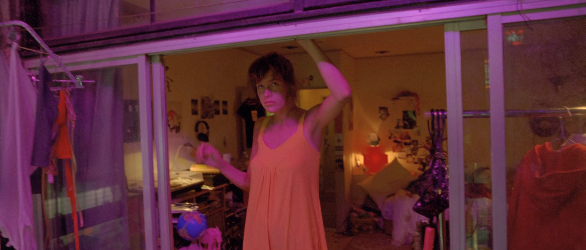 A woman in a pale orange slip stands with one arm raised in a cluttered room seen through glass, the space lit magenta and violet.