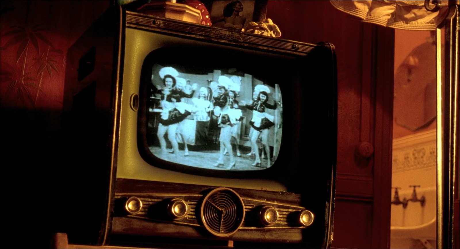 A television screen showing a black-and-white dance number, its cold blue-white light inside a hot amber room with warm practicals and a pale bathroom beyond an open door.