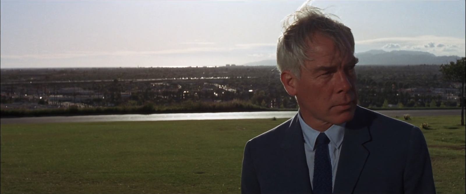 Lee Marvin in a dark suit and pale blue shirt on a lawn above a hazy Los Angeles basin, his grey hair catching the light.