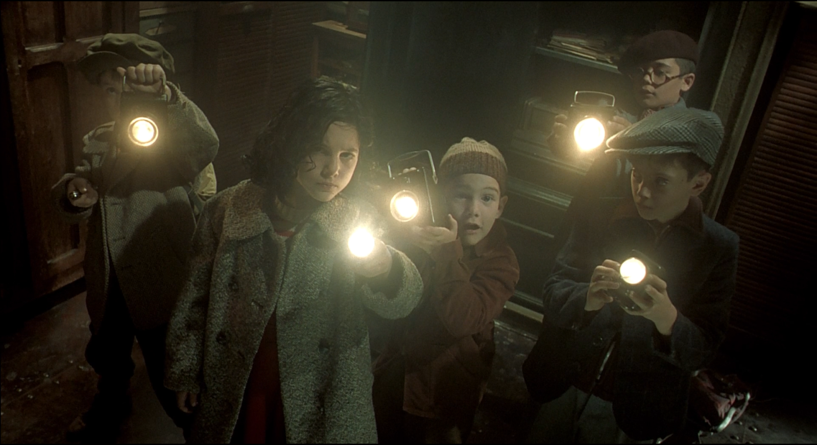 Five children in a dark interior, each holding a lantern up toward the camera, the edges of the frame in near-total darkness.