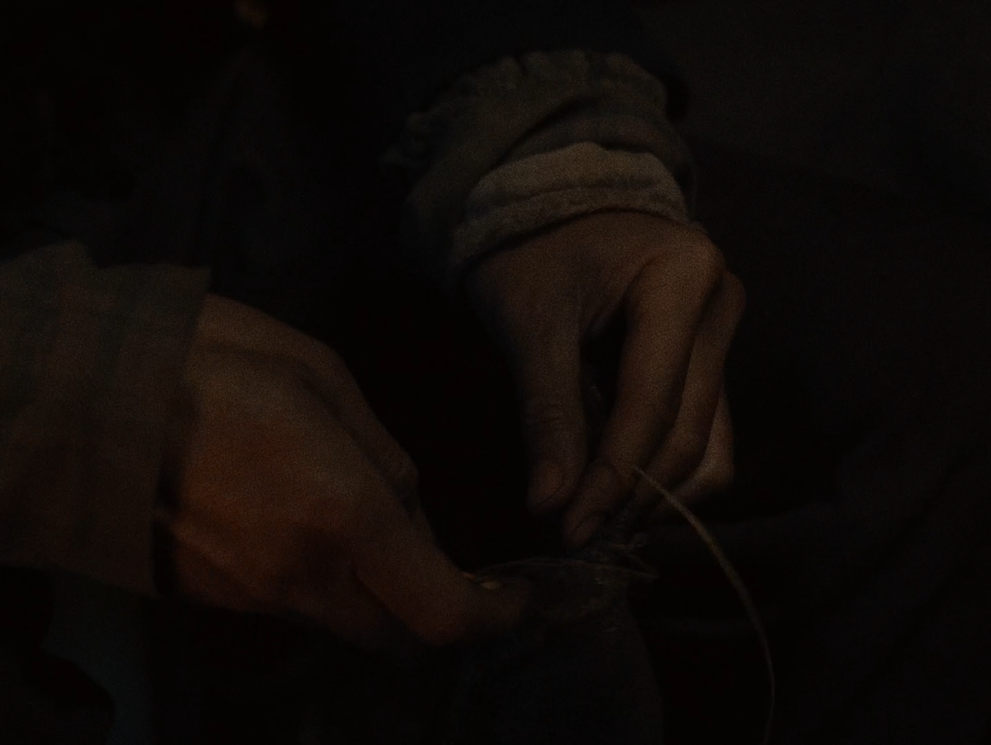 Two pairs of hands sewing by very low firelight, almost the entire frame in deep shadow.