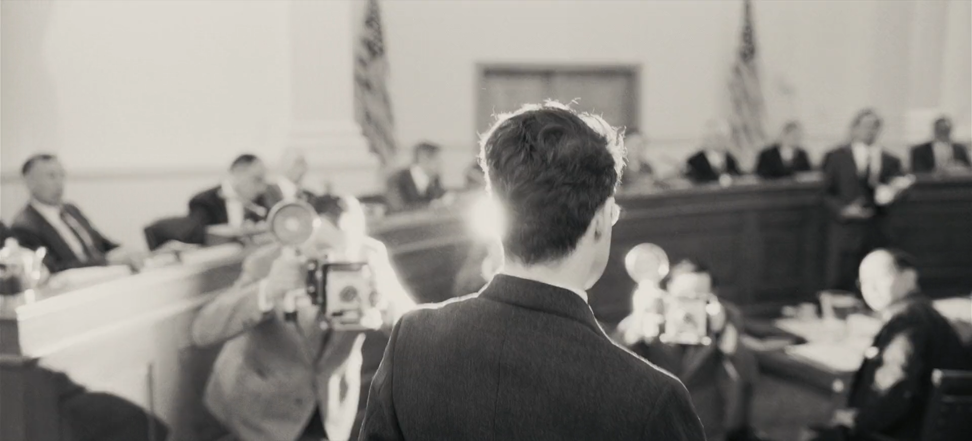 A black-and-white hearing room seen over a man's shoulder, press photographers' flashbulbs firing toward him beneath tall flags.