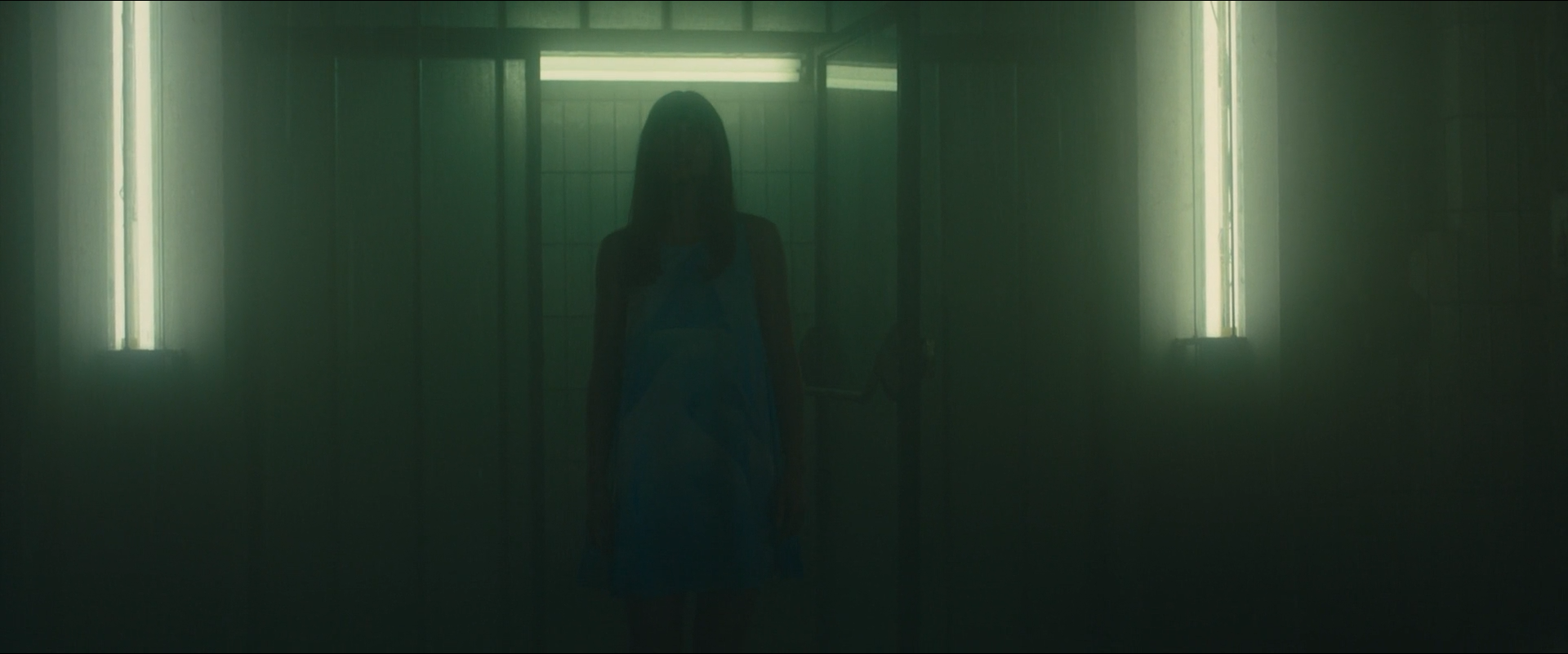 Shot 0227 from Blade Runner 2049 at 01:20:51:07. Two window slots and a lit doorway supply everything in a green-tinted corridor; the standing figure is lifted just enough to read as a pale blue shape without features.