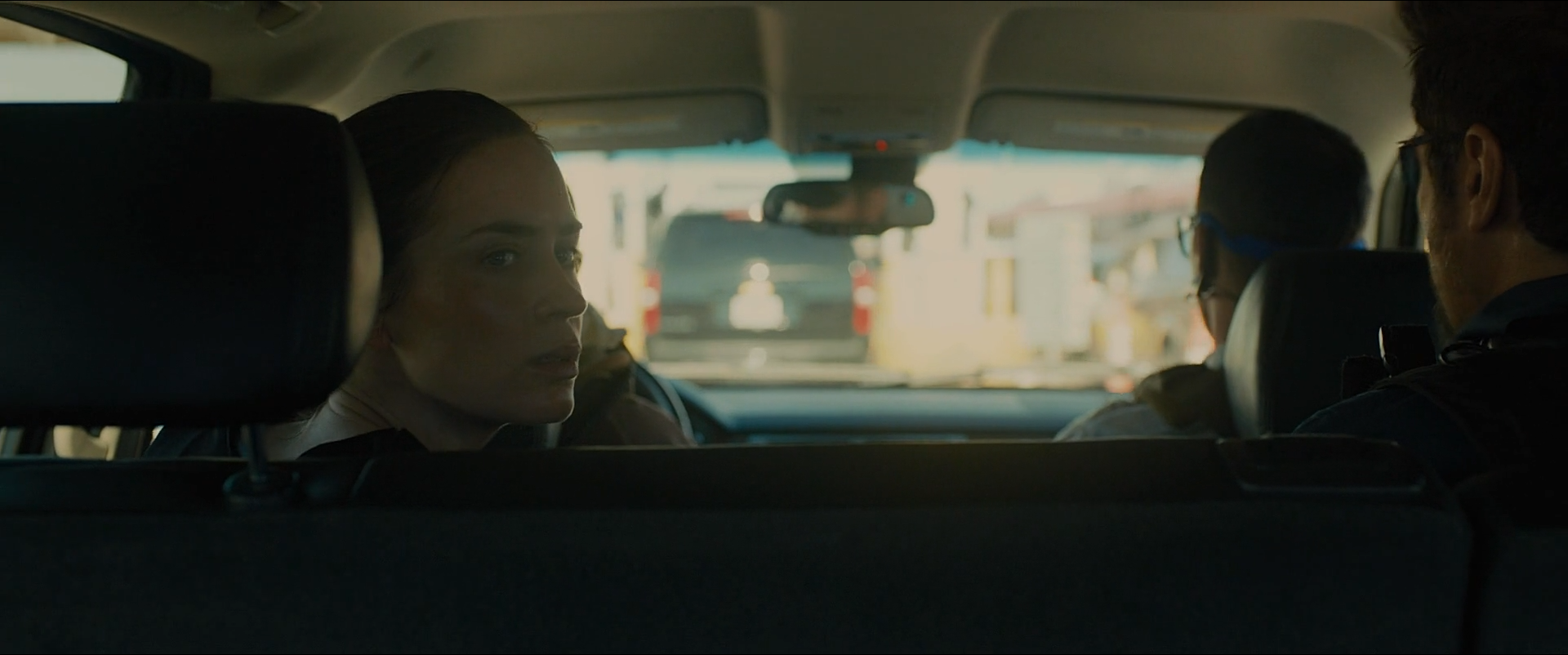 Shot 0346 from Sicario at 01:33:30:04. The car interior is held near black while the windscreen and side windows blow out to the crossing beyond; Kate's face catches just enough of that light to read against it.