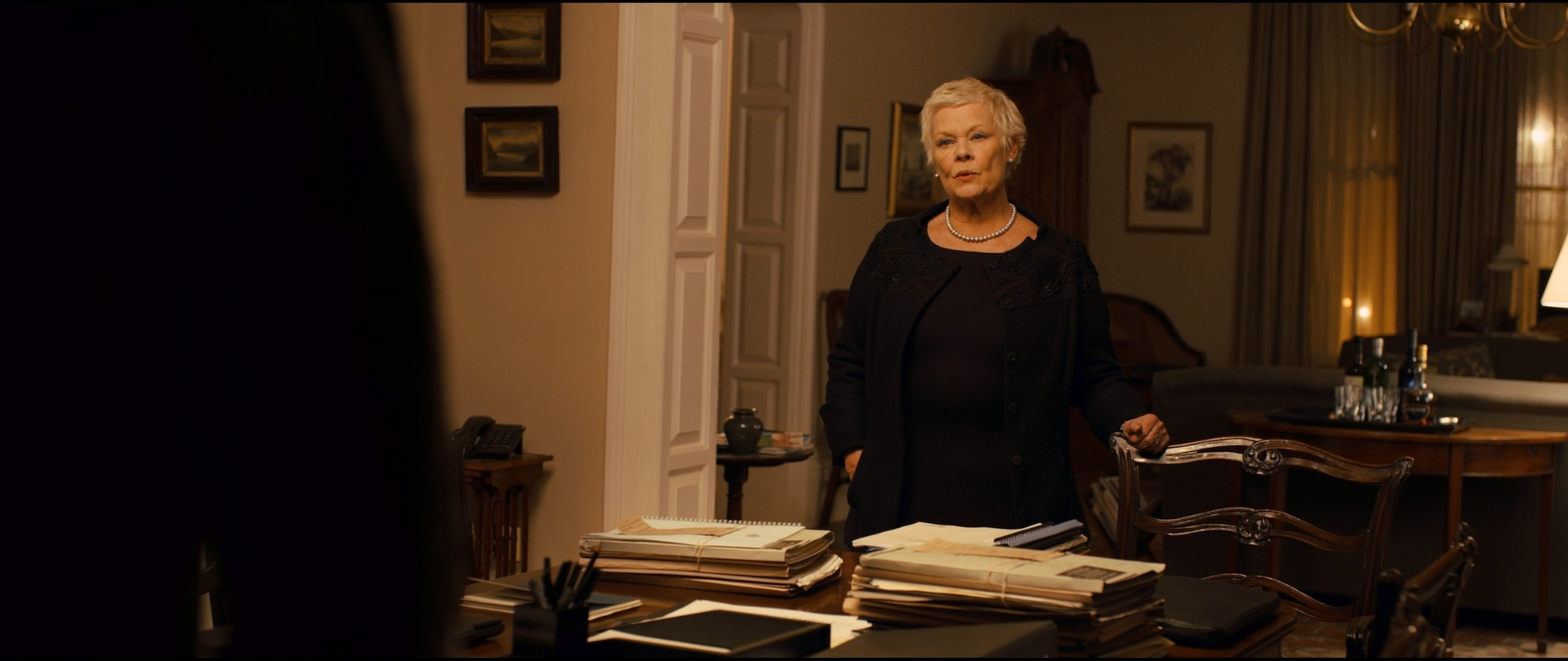 Shot 0648 from Skyfall at 01:27:14:20. Table lamps and a chandelier hold the room at a low amber level; M stands at centre in black while an out-of-focus foreground shoulder blocks the left of the frame to near-black.