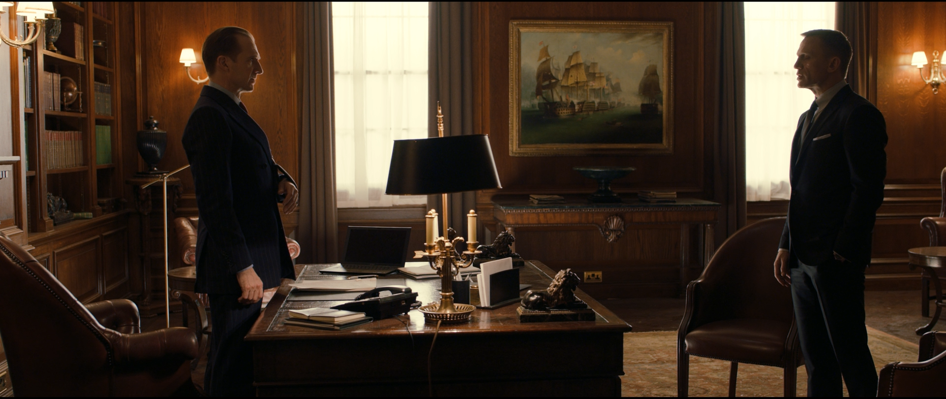 Shot 2619 from Skyfall at 03:17:31:15. Oak panelling, brass lamps and a naval painting set a warm, evenly lit room; the two dark suits stand at opposite edges of the frame with the desk and window between them.