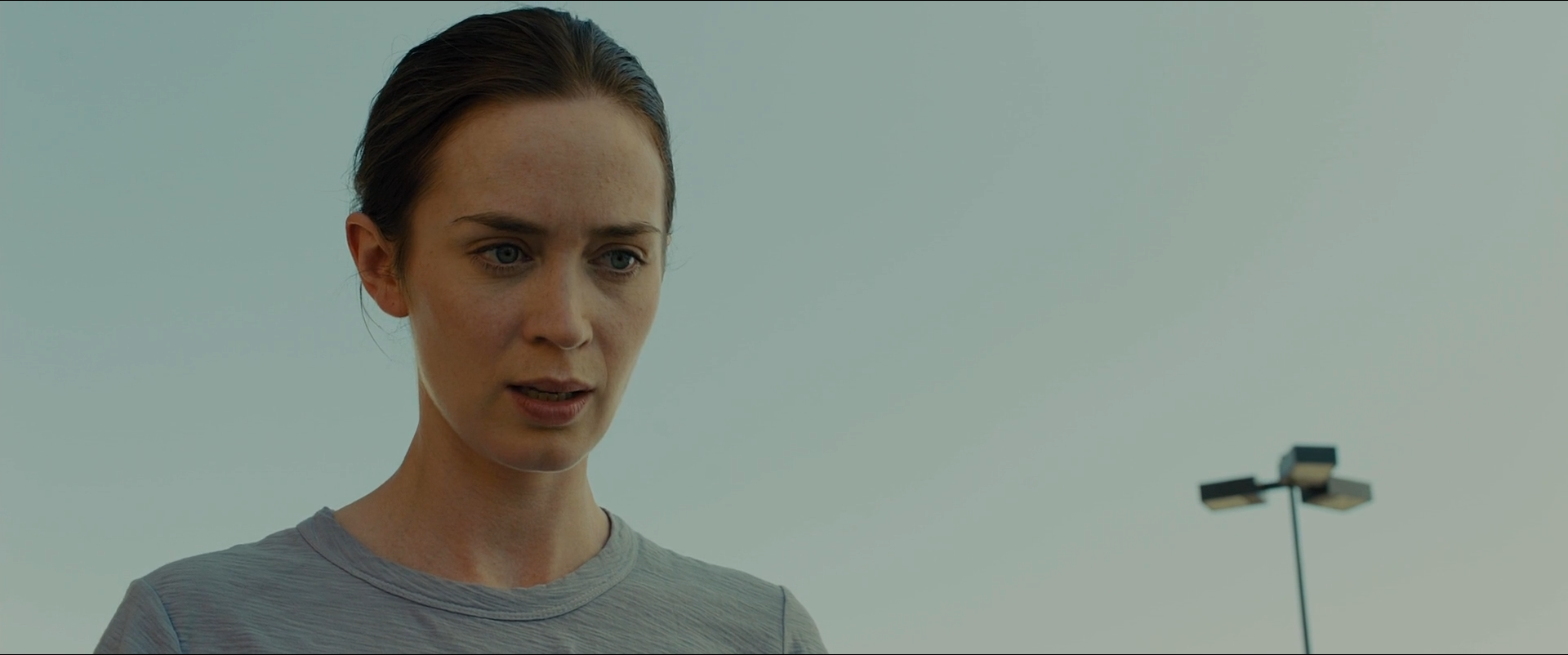 Shot 0590 from Sicario at 01:59:42:04. An overcast sky fills the background at a flat pale grey; Kate's face and grey shirt sit only slightly below it, and a distant lamp standard is the only other mark.