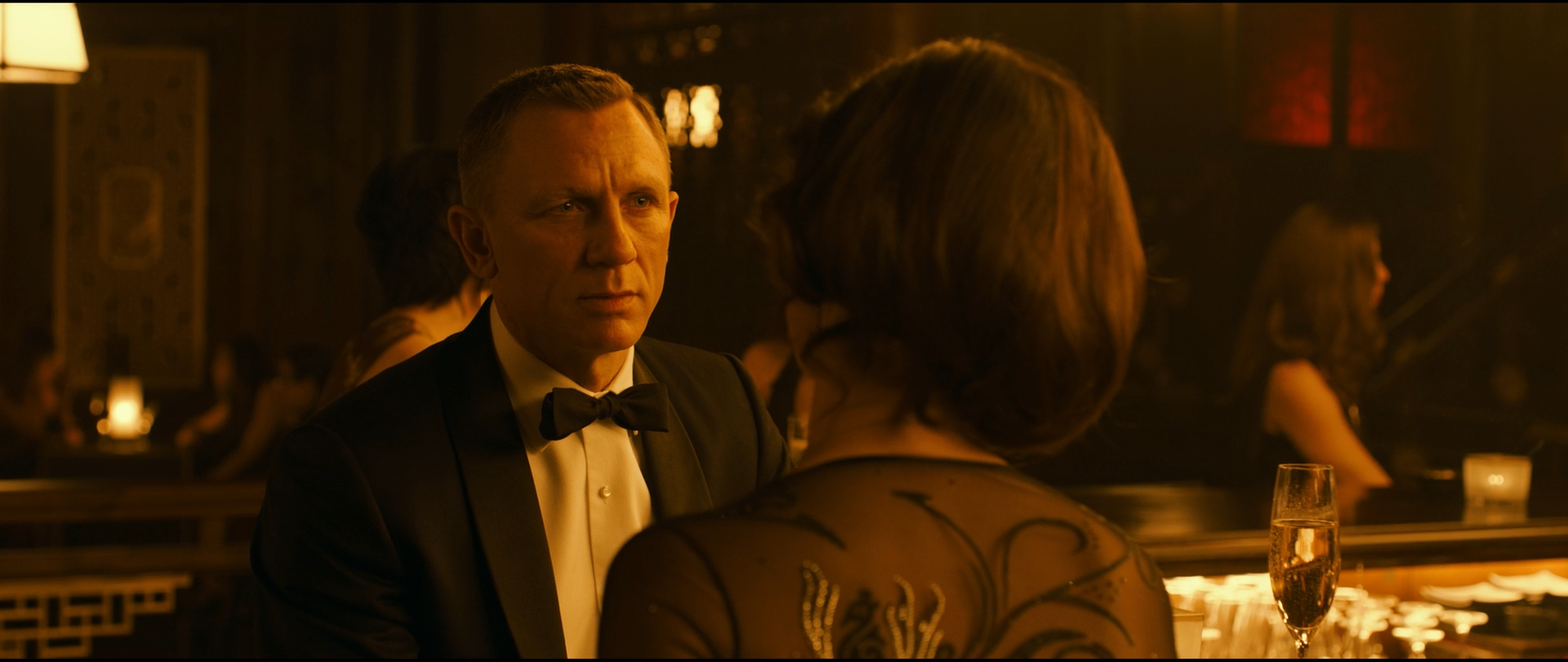 Shot 1100 from Skyfall at 02:00:54:23. Practical lamps and gilded screens keep the room in one amber register; the black dinner jacket, white shirt and the woman's dark embroidered back are the only strong tonal breaks.