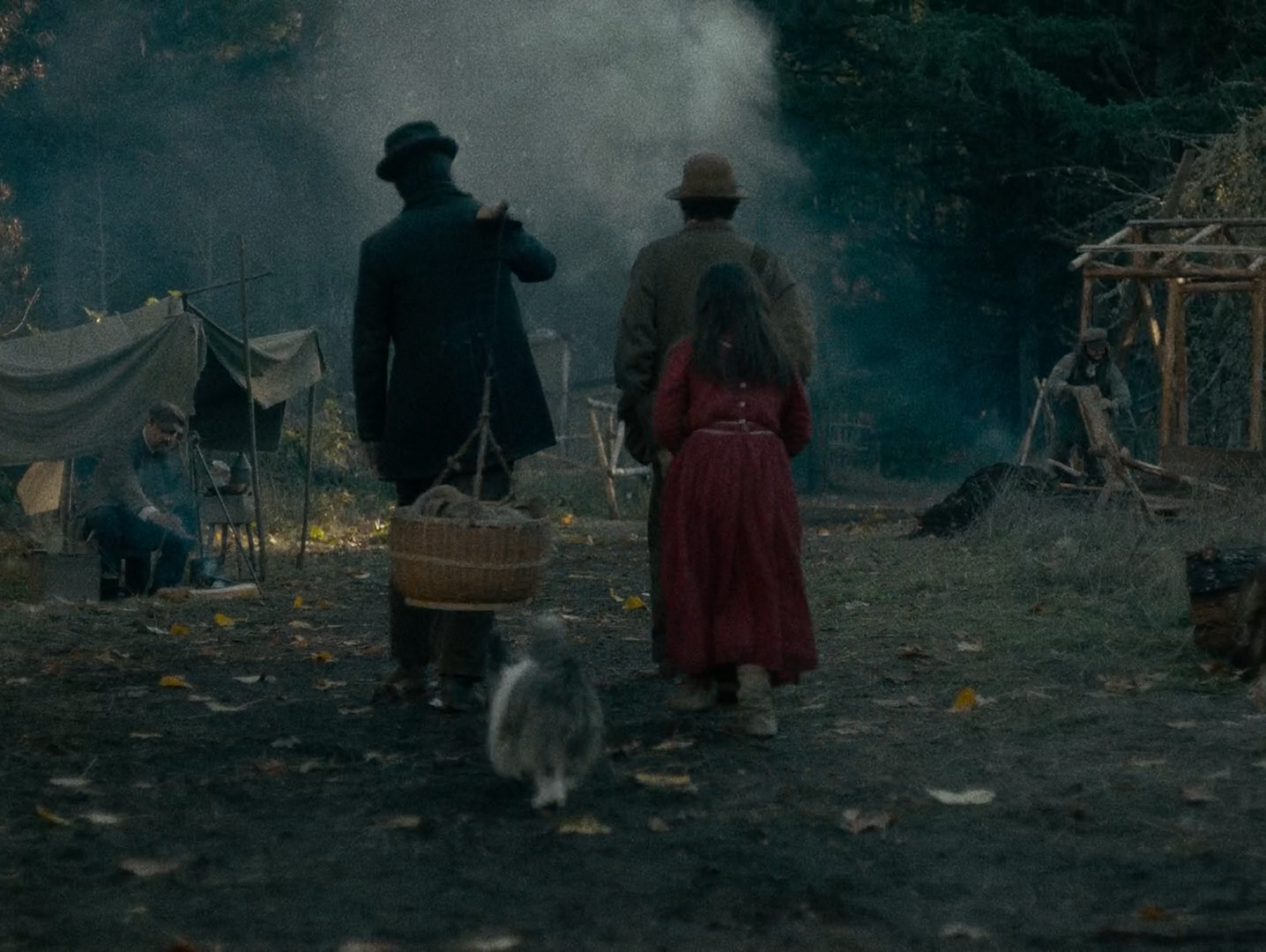 Seen from behind, two men and a girl in a red dress walk away through a woodland camp with canvas shelters and a fire, a white dog beside them and smoke hanging among the trees.