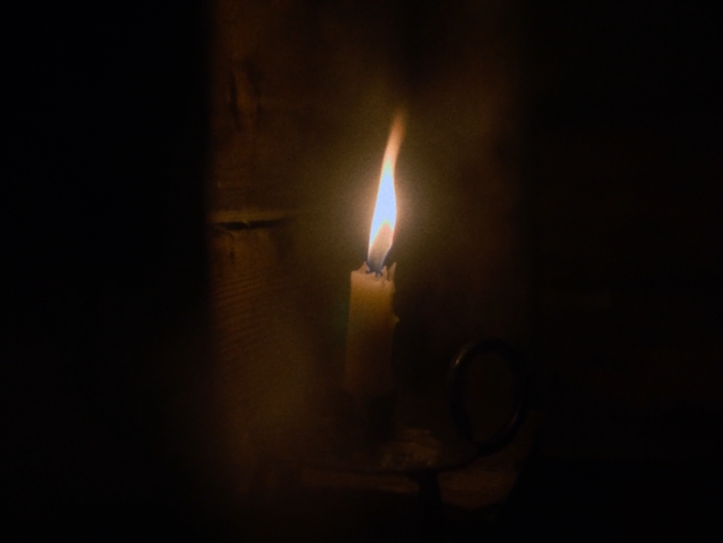 A lit candle in a dark iron holder stands against rough vertical timber boards, its flame the only light in an otherwise black frame.