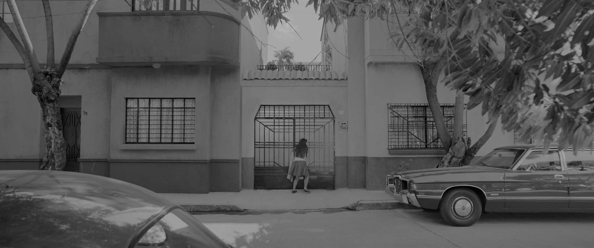 A residential street facade in flat daylight. An upper storey with a curved balcony rises at the left, a barred window with net curtains sits below it, a tall barred gate stands at the centre and another barred window at the right. A band of darker paint runs along the base of the wall. A small girl in a pale sleeveless top and pale skirt stands with her back to the camera at the gate. A large pale saloon is parked at the right, the roof of another car crosses the bottom left corner, and two street trees rise through the frame with a bough of dark leaves across the top right.