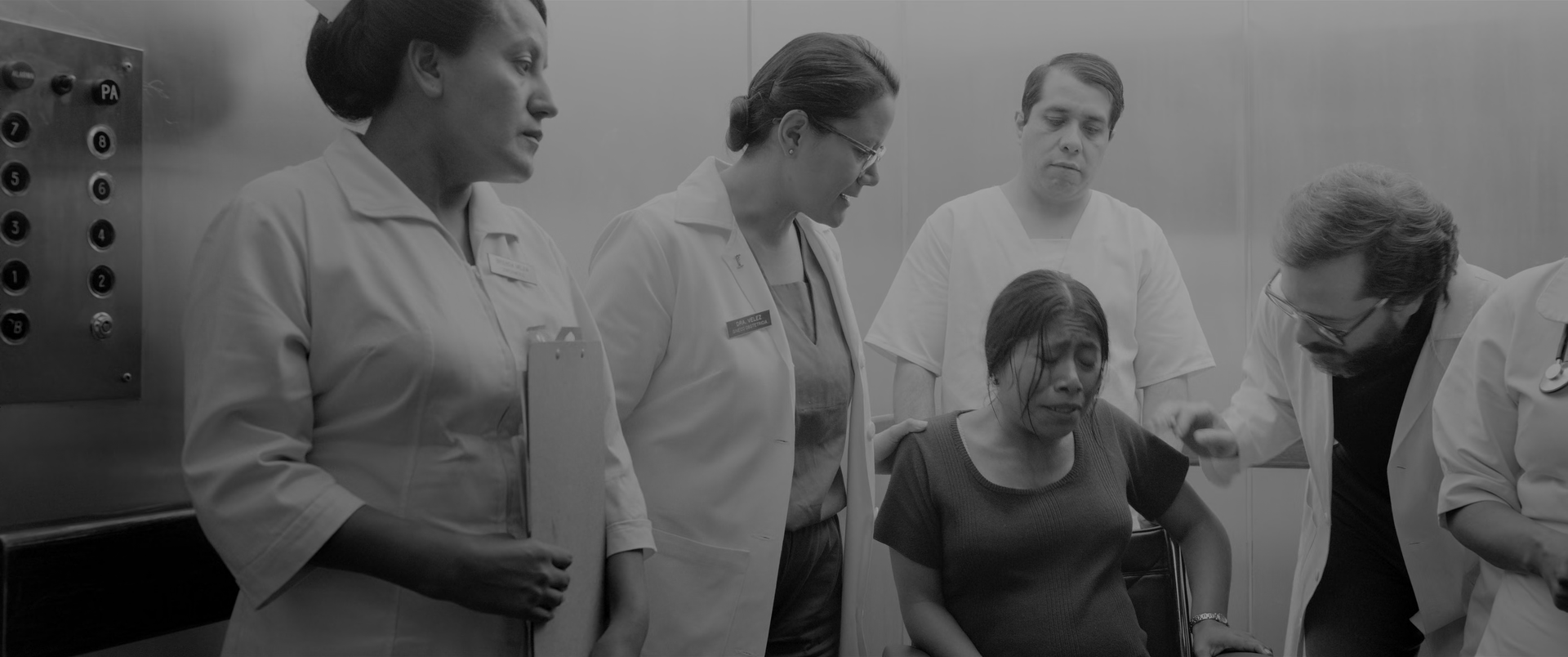 Inside a hospital lift. A nurse in a white uniform stands at the left holding a clipboard, a doctor in a white coat and glasses beside her, an orderly in white scrubs behind, and a second doctor in a white coat leans in from the right. A woman in a dark top sits in a wheelchair at the centre right, her face lowered and crumpled in distress. A lift button panel is at the far left and pale wall panels close the background.