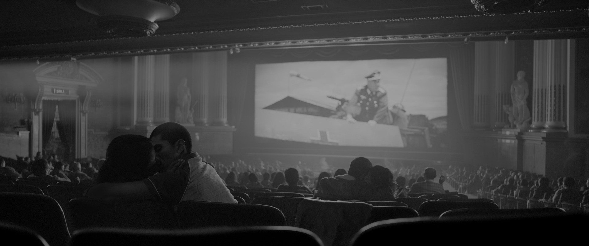 A cinema auditorium photographed from behind the audience. A large lit screen at the centre right shows a man in a peaked military cap in a boat. Ornate plasterwork, pilasters and statues line the walls, a haze of projector light crosses the upper frame, and rows of dark seat backs and heads fill the lower half. A couple at the centre left lean together, their faces almost touching, lit only by the screen.