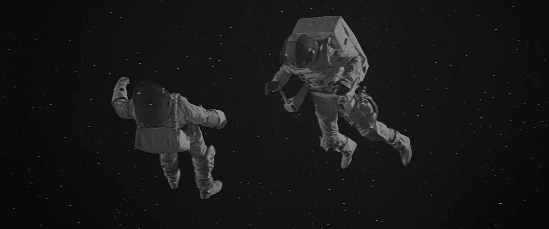 Two figures in bulky white spacesuits with dark visored helmets drift against a black starfield that fills the whole frame. The left figure is turned away with one arm raised; the right figure carries a large angular box-shaped pack and holds a tool. Small white points of light are scattered across the black background.