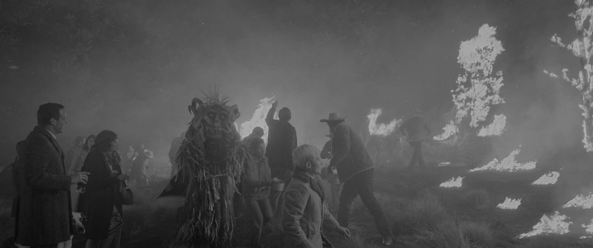 A night scene in heavy smoke. At the left, guests in overcoats hold drinks; at the centre left a figure in a shaggy full-body costume with a horned mask stands facing the camera. A man in a wide-brimmed hat swings a container toward the fire at the centre right, a fair-haired child in a coat walks through the foreground, and burning trees and patches of ground fire fill the right of the frame behind a veil of smoke.