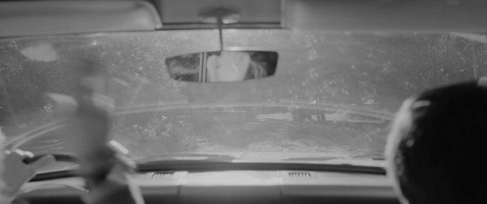The view forward from inside a car. A dusty, scratched windscreen fills most of the frame, scattering light into a bright veil, with trees and a road faintly visible through it. A rear-view mirror hangs at the top centre with a face reflected in it. A blurred hand and arm cross the lower left, a wiper arm lies along the bottom of the glass, and a dark head in silhouette occupies the right edge.