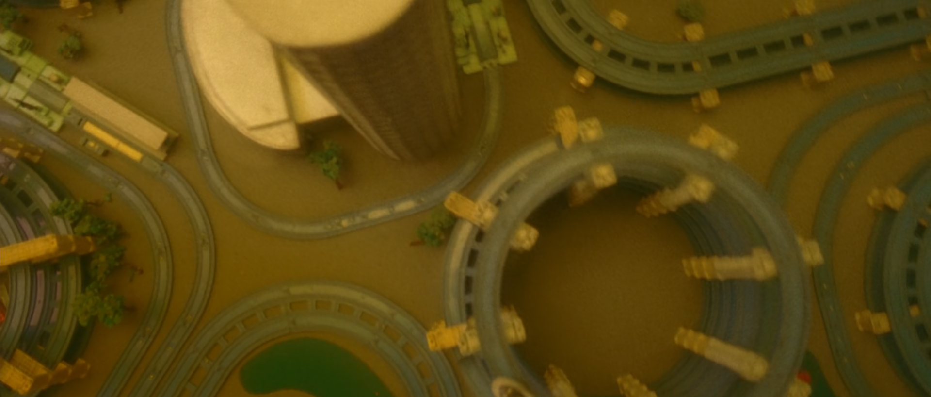 A directly overhead view of a city intersection with curved elevated roads, rounded structures, small vehicles and trees, all rendered in muted yellow-green and brown.