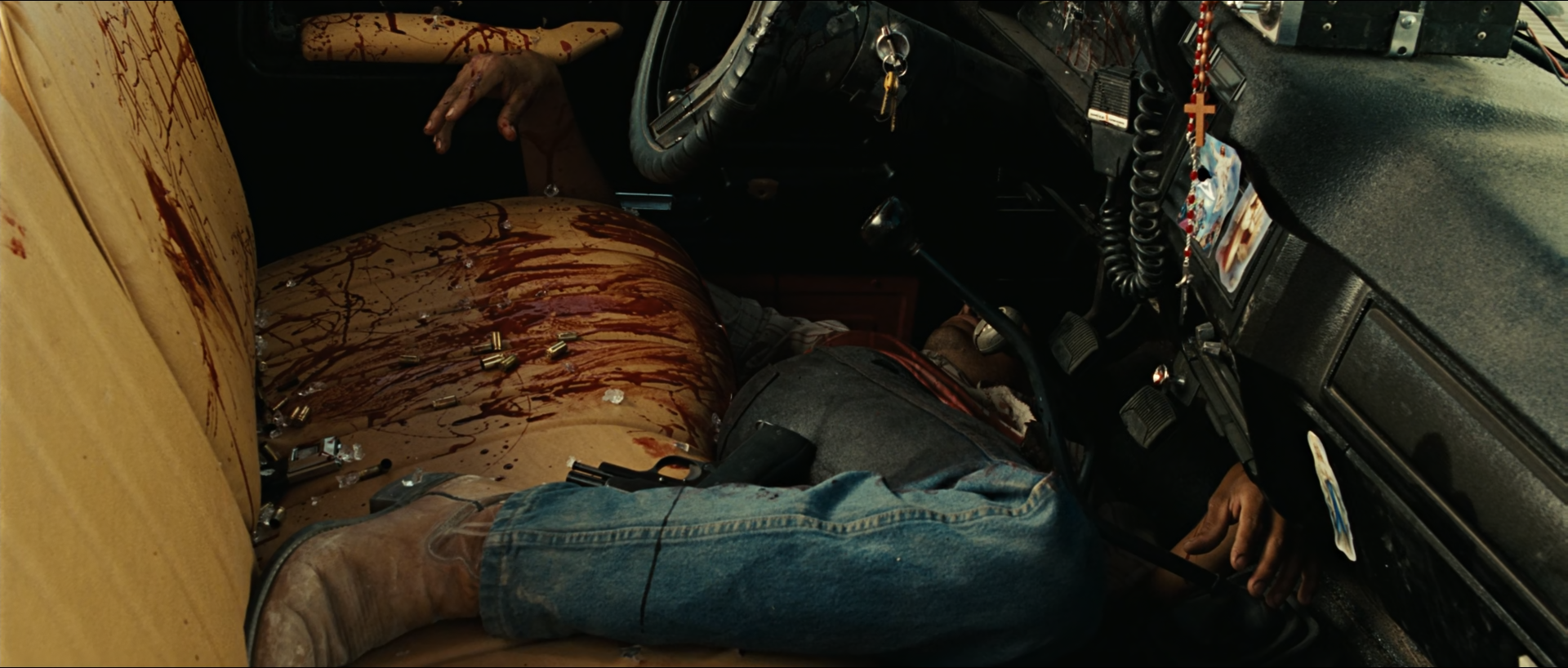 Inside a pickup cab, a man's body in a bloodied shirt and blue jeans lies across a cream vinyl seat streaked with dark red blood and scattered brass casings, beside a black dashboard hung with a rosary.