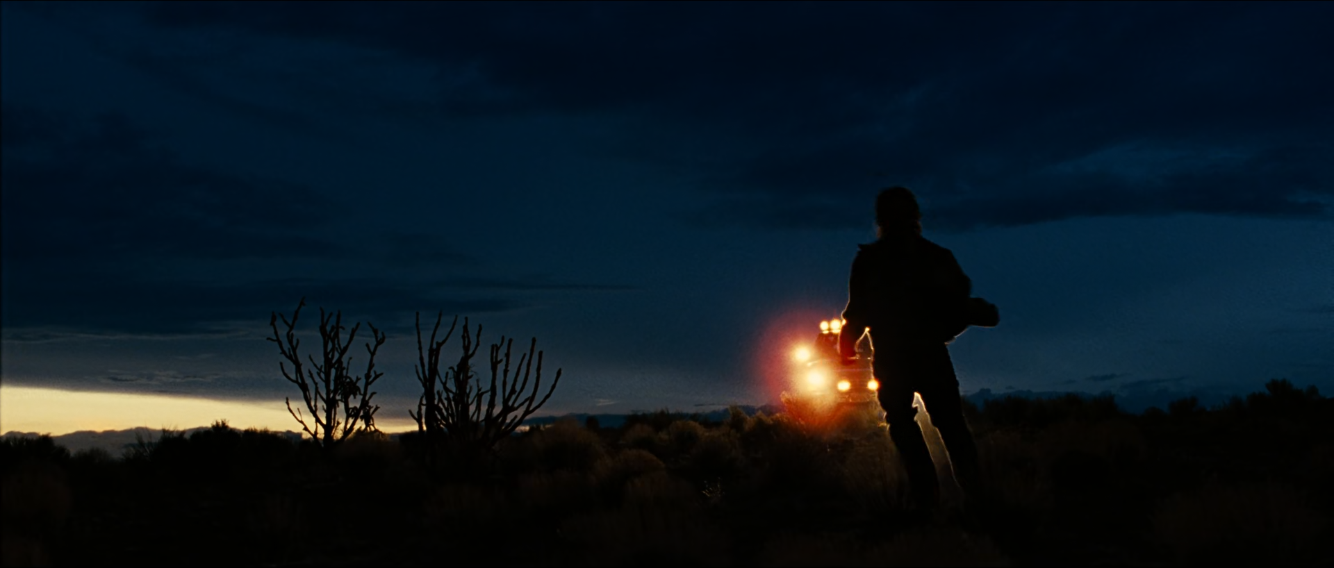 At dusk in open desert, a silhouetted man carrying something runs across dark scrub while a vehicle's amber headlights glow behind him under a deep blue cloudy sky with a faint orange band on the horizon.
