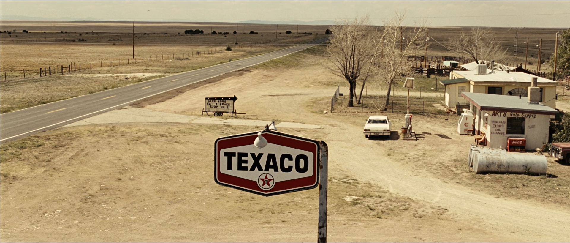 An elevated view of a desert filling station: a red and white Texaco sign in the foreground, a beige dirt lot, a two-lane road, a white car, low buildings and bare trees under a pale sky.
