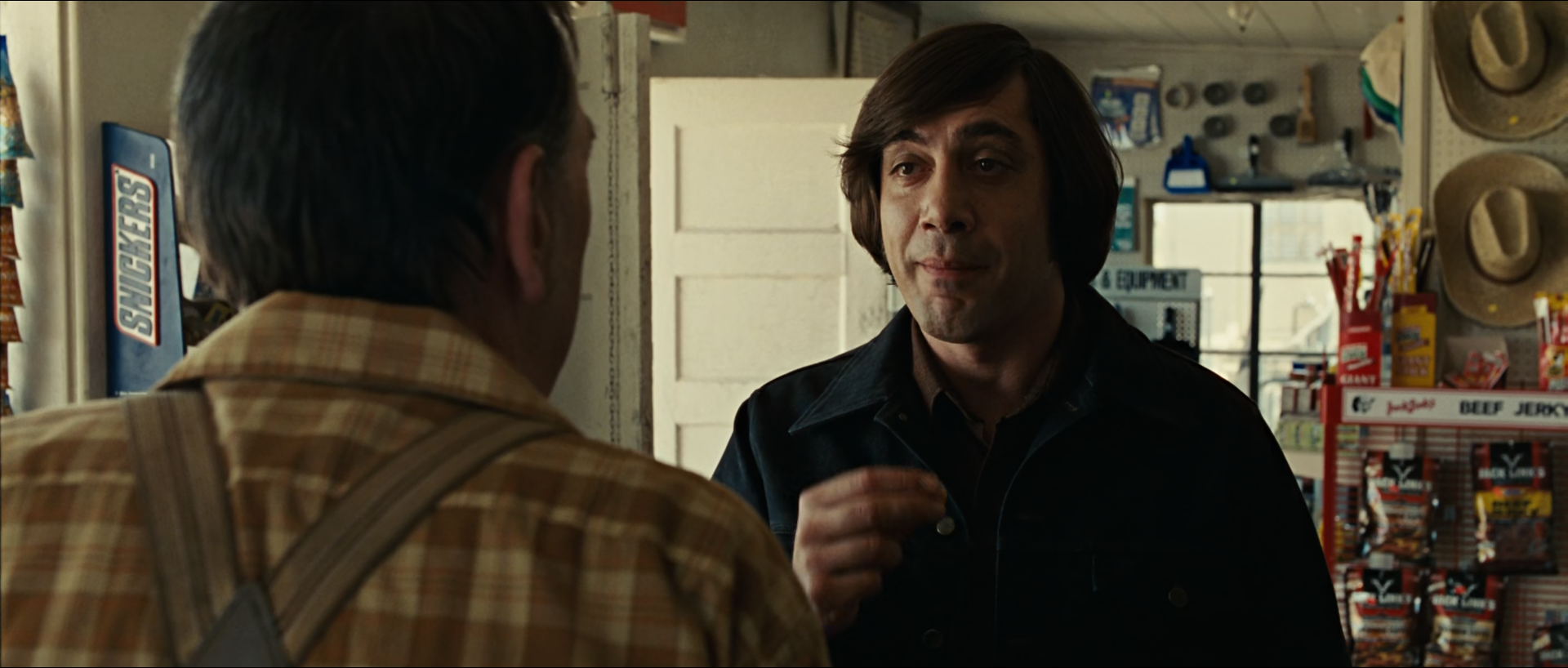 Inside a small store, a man with a dark bowl haircut and black jacket stands behind a counter facing an older man seen from behind in a plaid shirt and suspenders, with a white door, hats and snacks in the background.