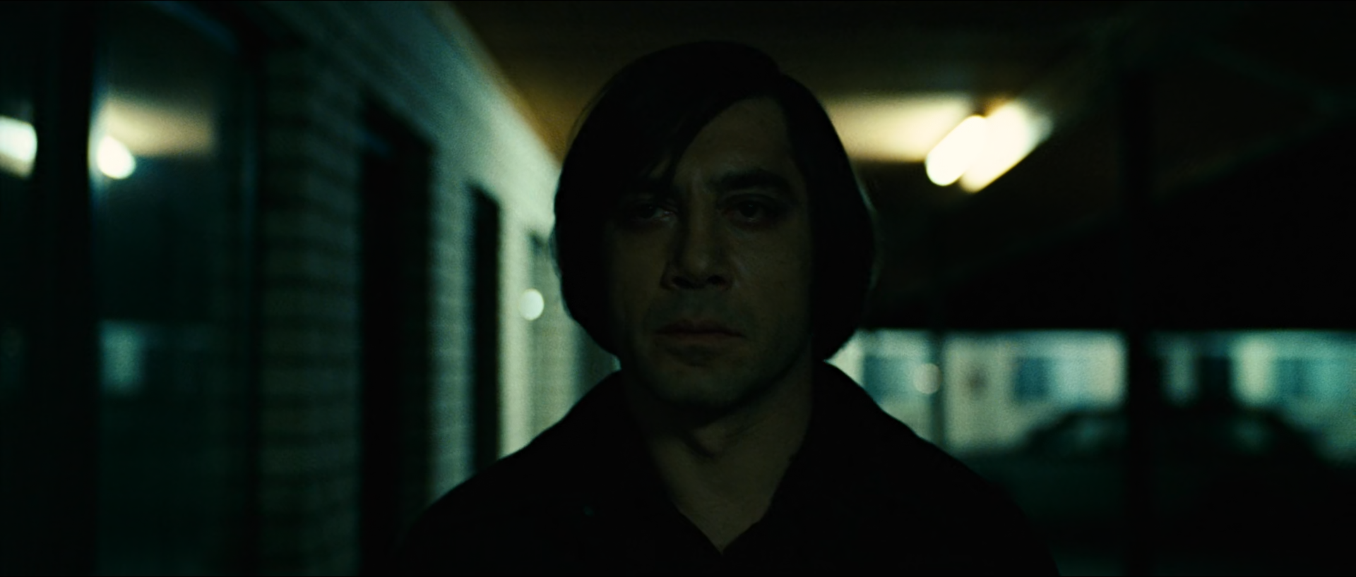 A man with a dark bowl haircut and black jacket walks toward the camera along a dim motel corridor at night, lit by soft green-white fluorescent tubes behind him and brick walls in dark green shadow.