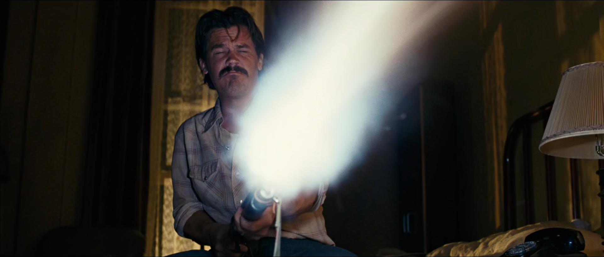 In a dim brown motel room, a man with a moustache squints with eyes shut as a broad bright white-blue burst of light extends diagonally from the weapon he holds, with a table lamp and bedframe beside him.