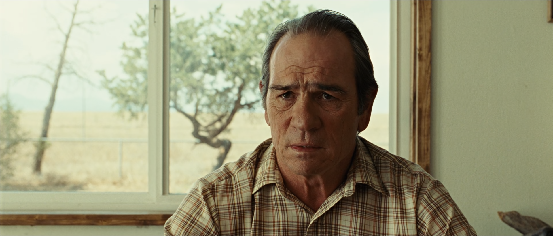 An older man with grey hair and a lined face in a brown plaid shirt sits facing the camera in a pale room, with a bright window behind him showing a sunlit dry field and a crooked tree.