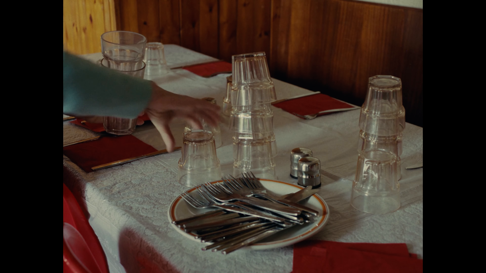 A hand places glassware above a pale tablecloth with red napkins and metal cutlery; creases and reflections distinguish the light surfaces.