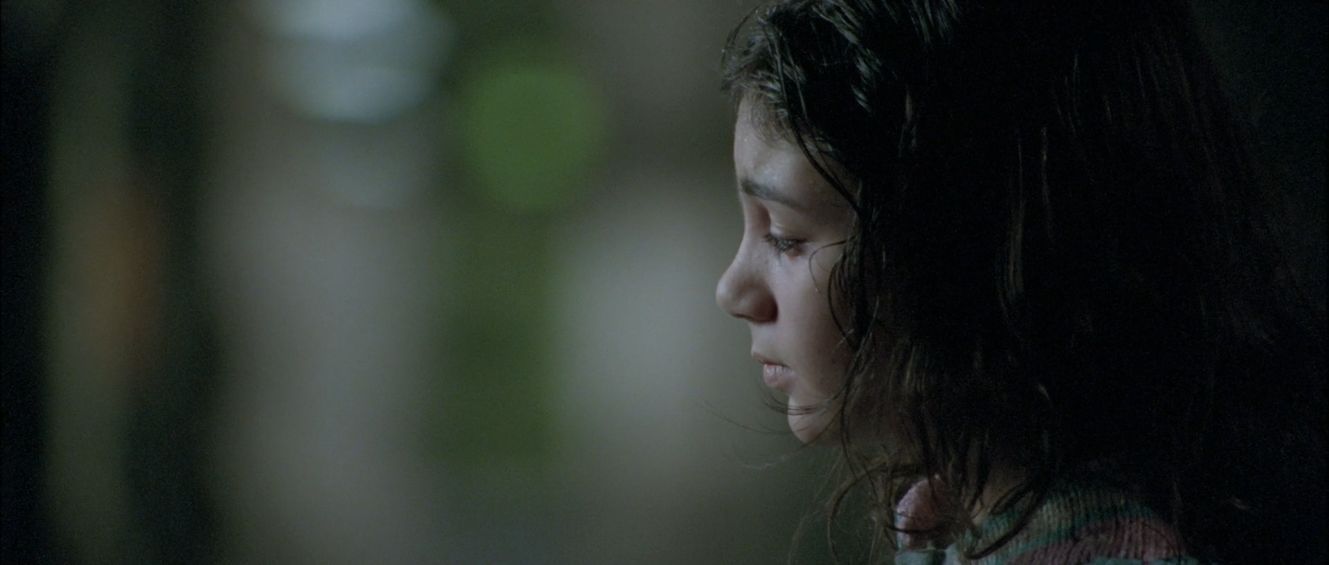 A dark-haired girl appears in right-facing profile, softly lit on her cheek and nose against a blurred dark green background.