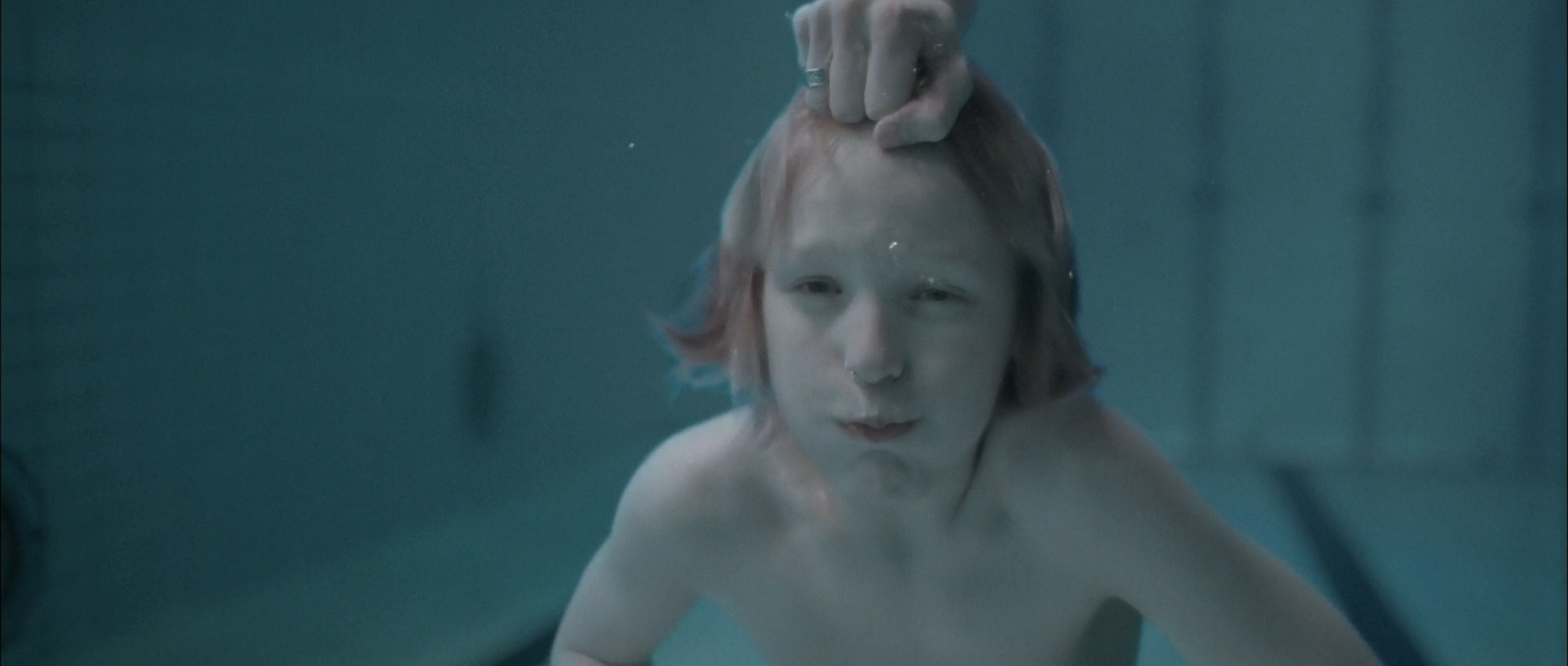 Underwater, a pale boy faces the camera while a hand grips the hair at the top of his head, with bubbles and a cyan pool wall around him.