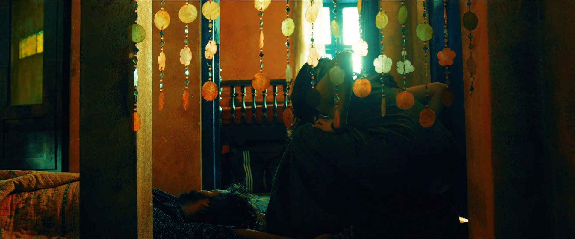 Dark figures stand behind hanging beads or shells, with orange walls and a bright window edged in teal beyond them.