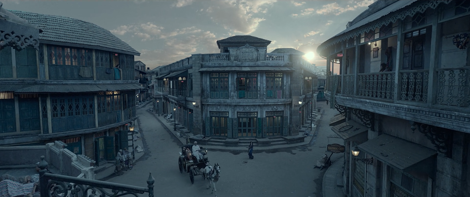 A high wide view over a period city crossroads at blue-grey dawn or dusk, with tiny warm lights and a low sun at the right.