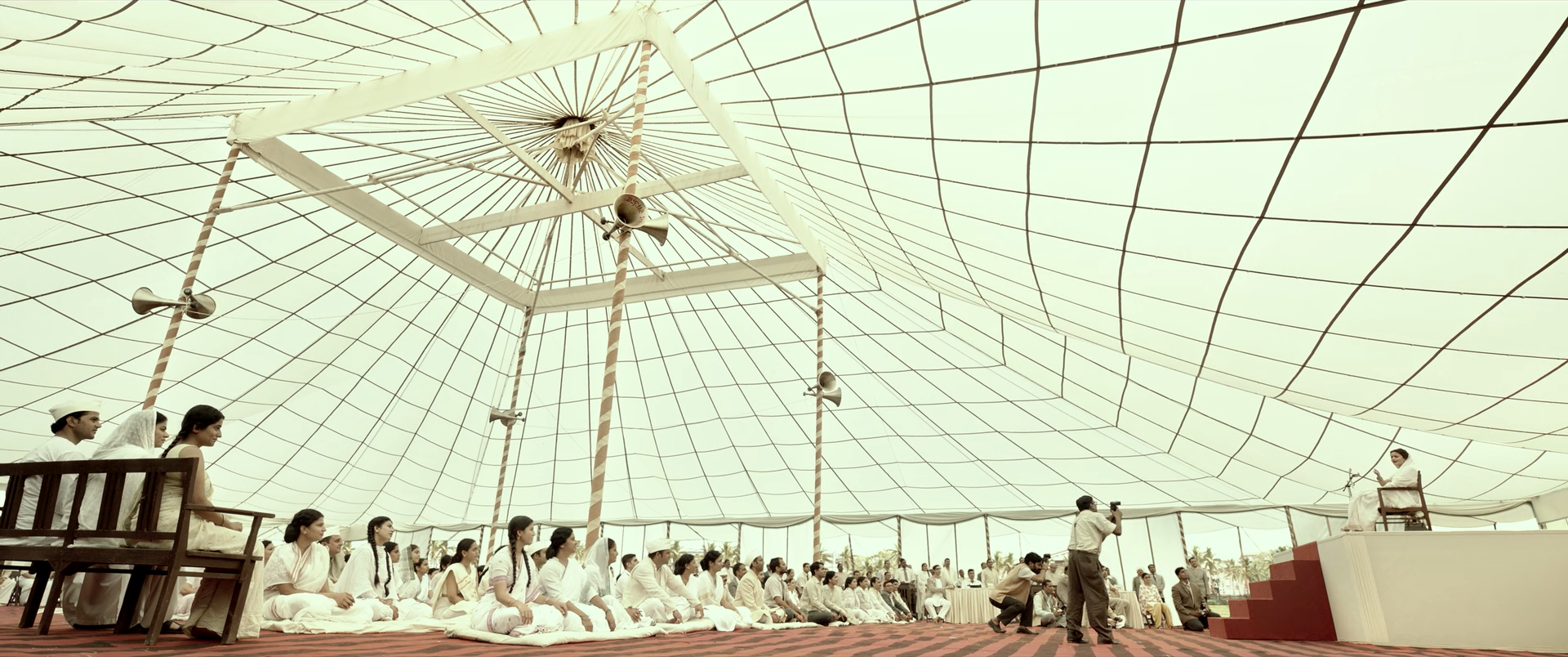 A vast crowd dressed in white gathers beneath an enormous cream-white tent canopy above a red and black patterned floor.